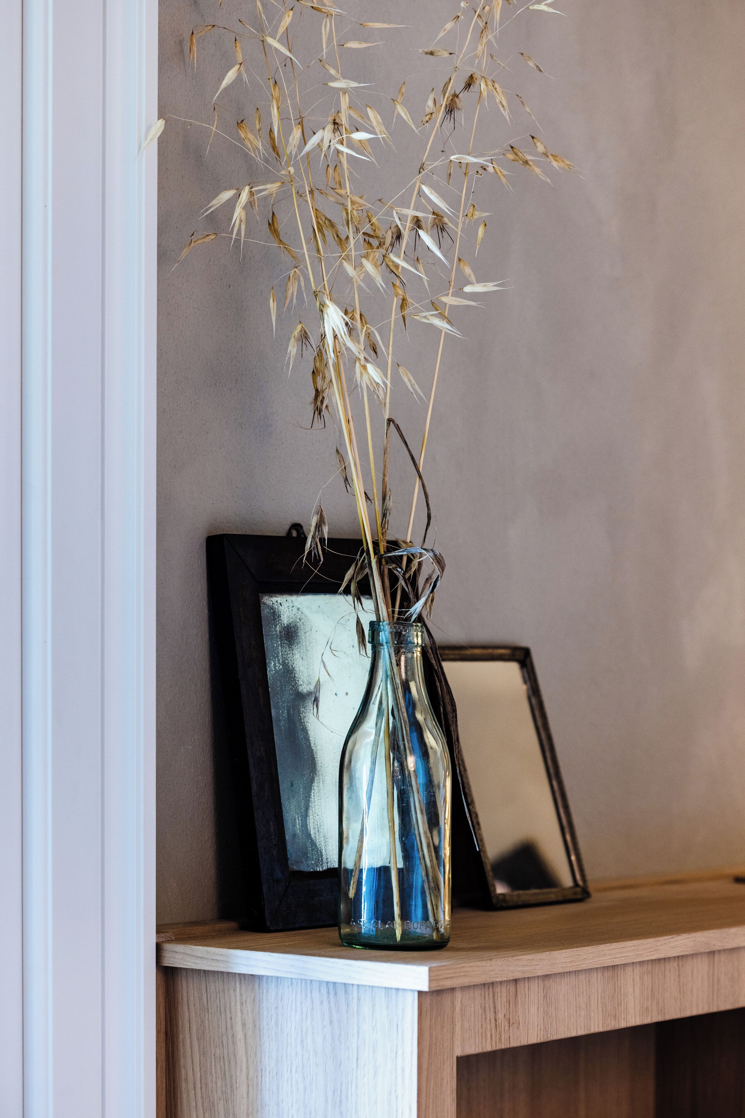 A clear glass vase with dried plants on a wooden shelf against a plain wall.
