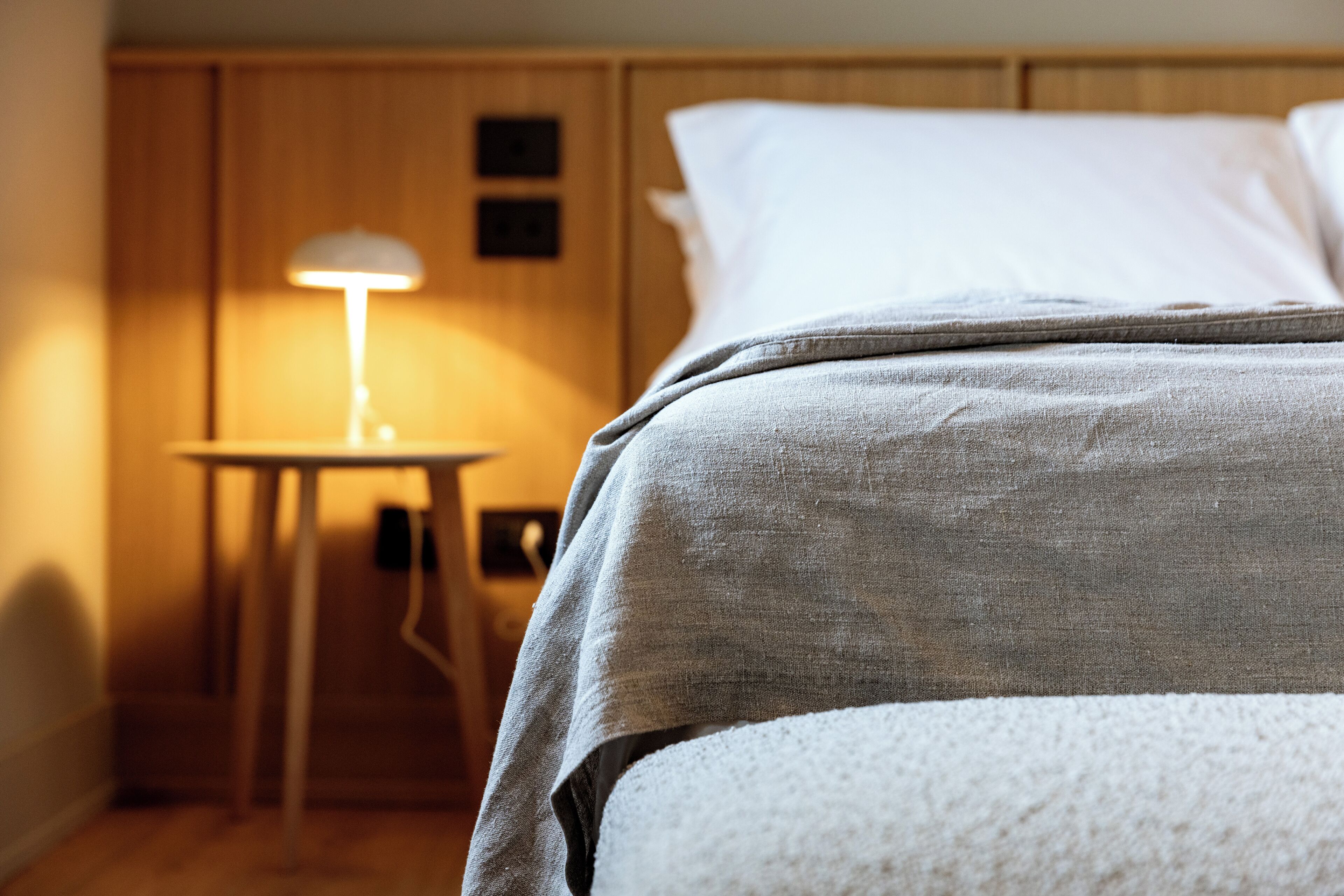 A neatly made bed with a grey blanket, a bedside lamp, and a wooden nightstand.
