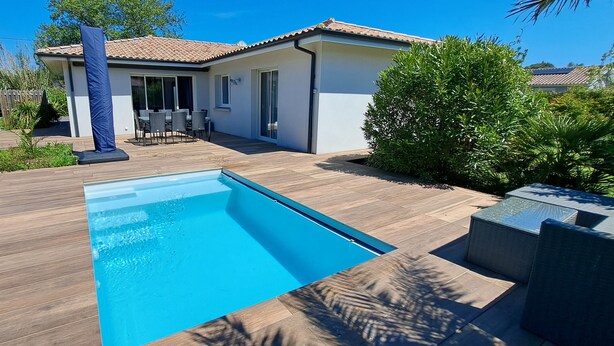 Una piscina amb una terrassa de fusta, una zona de pati amb mobles d'exterior, i una casa amb teulada enrajolada.