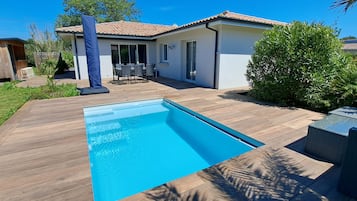 A swimming pool with a wooden deck, a patio area with outdoor furniture, and a house with a tiled roof.