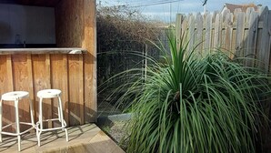 Un pati amb paviment de fusta, dos tamborets blancs, una tanca de fusta i una planta verda gran.