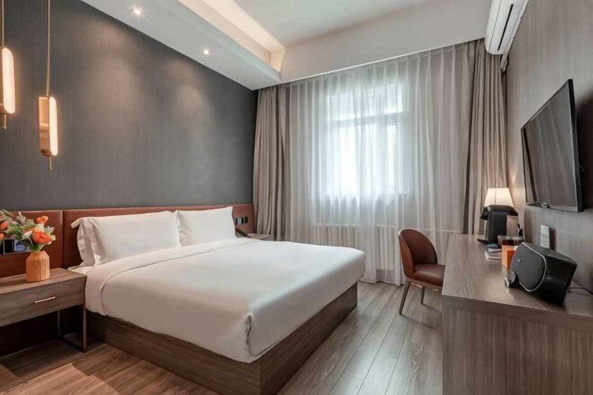 A modern hotel room with a large bed, a desk with a laptop, a TV, and a window with curtains.