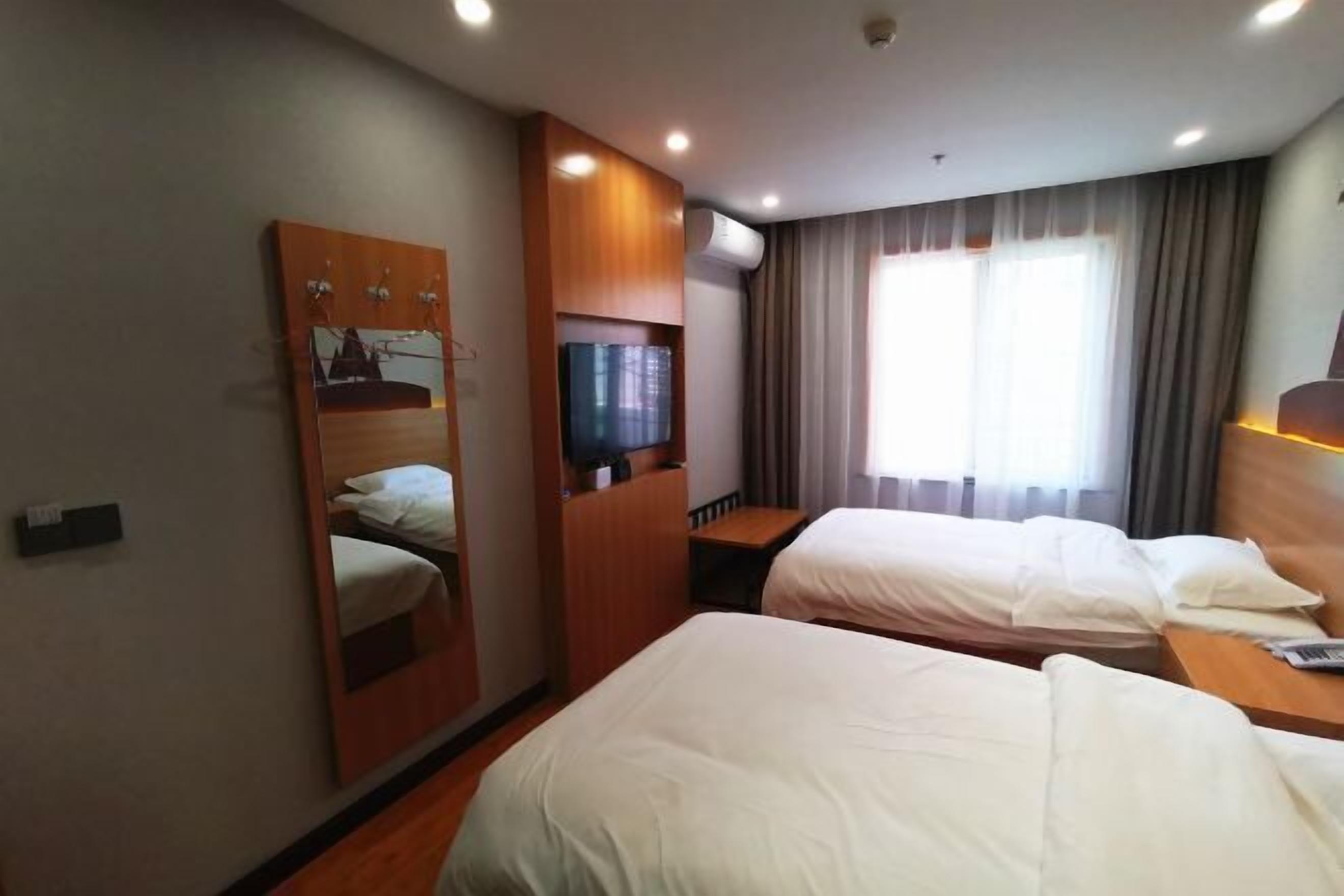 A hotel room with two beds, a TV, a desk, and a large window with curtains.