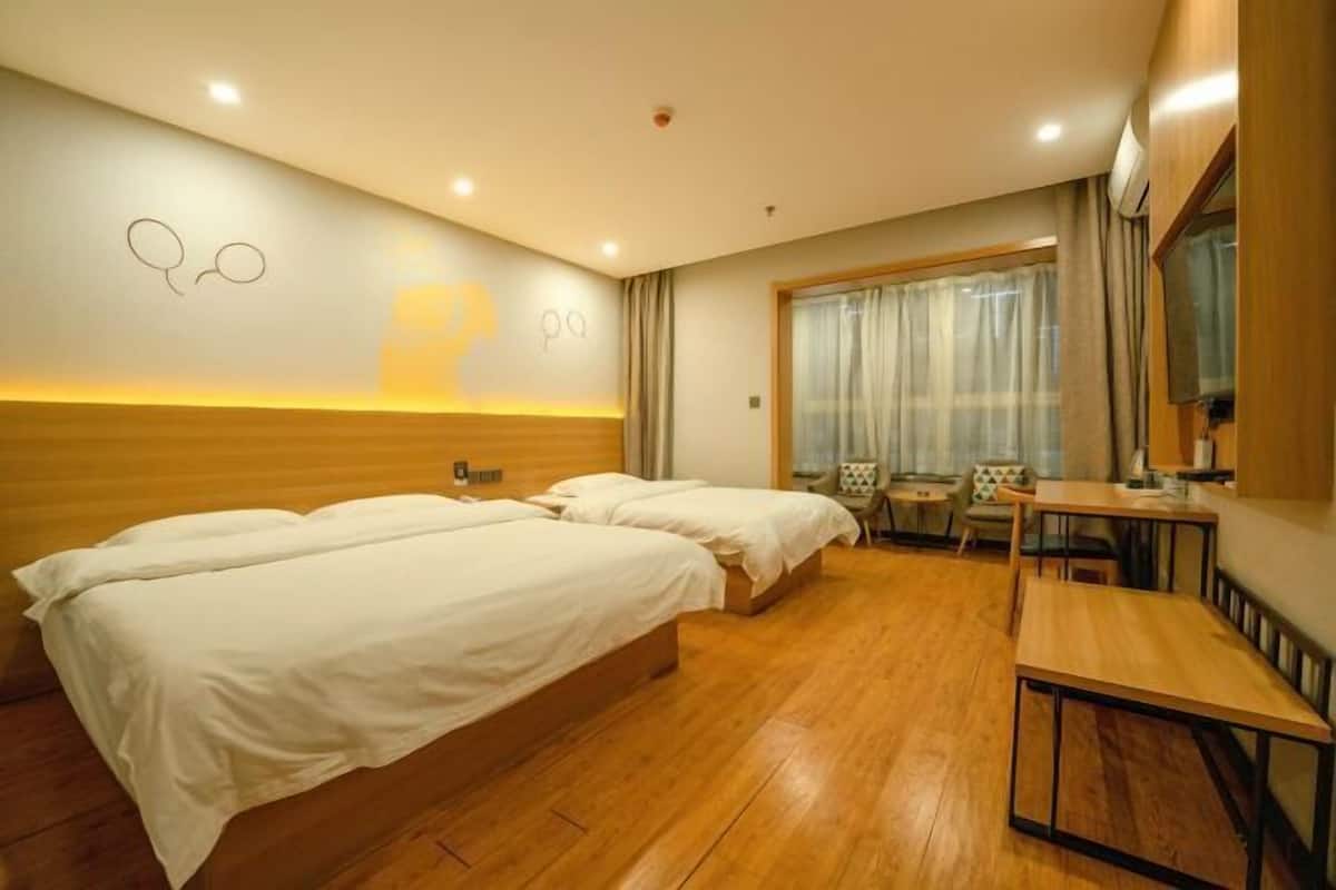 Habitación de hotel con dos camas, piso de madera, un escritorio, una silla, ventana con cortinas y una lámpara de pared junto a una obra de arte.