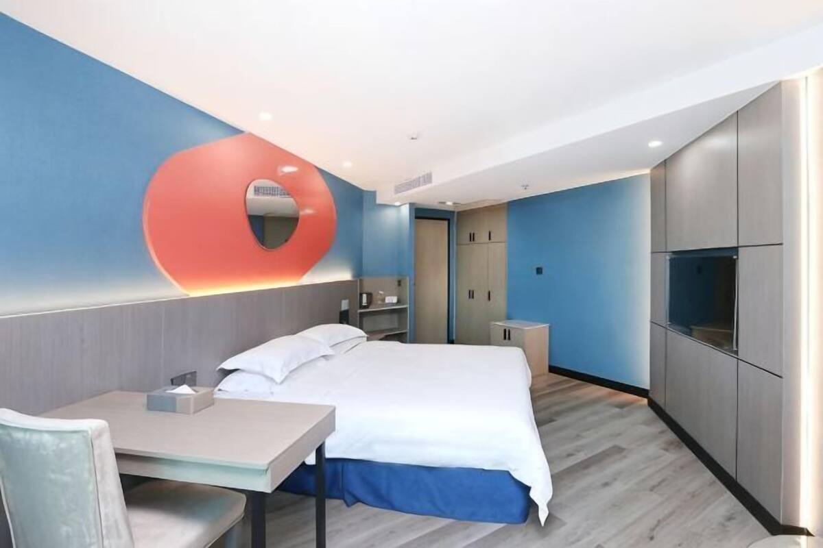 Quarto de hotel com cama, escrivaninha, cadeira, televisão e guarda-roupa.