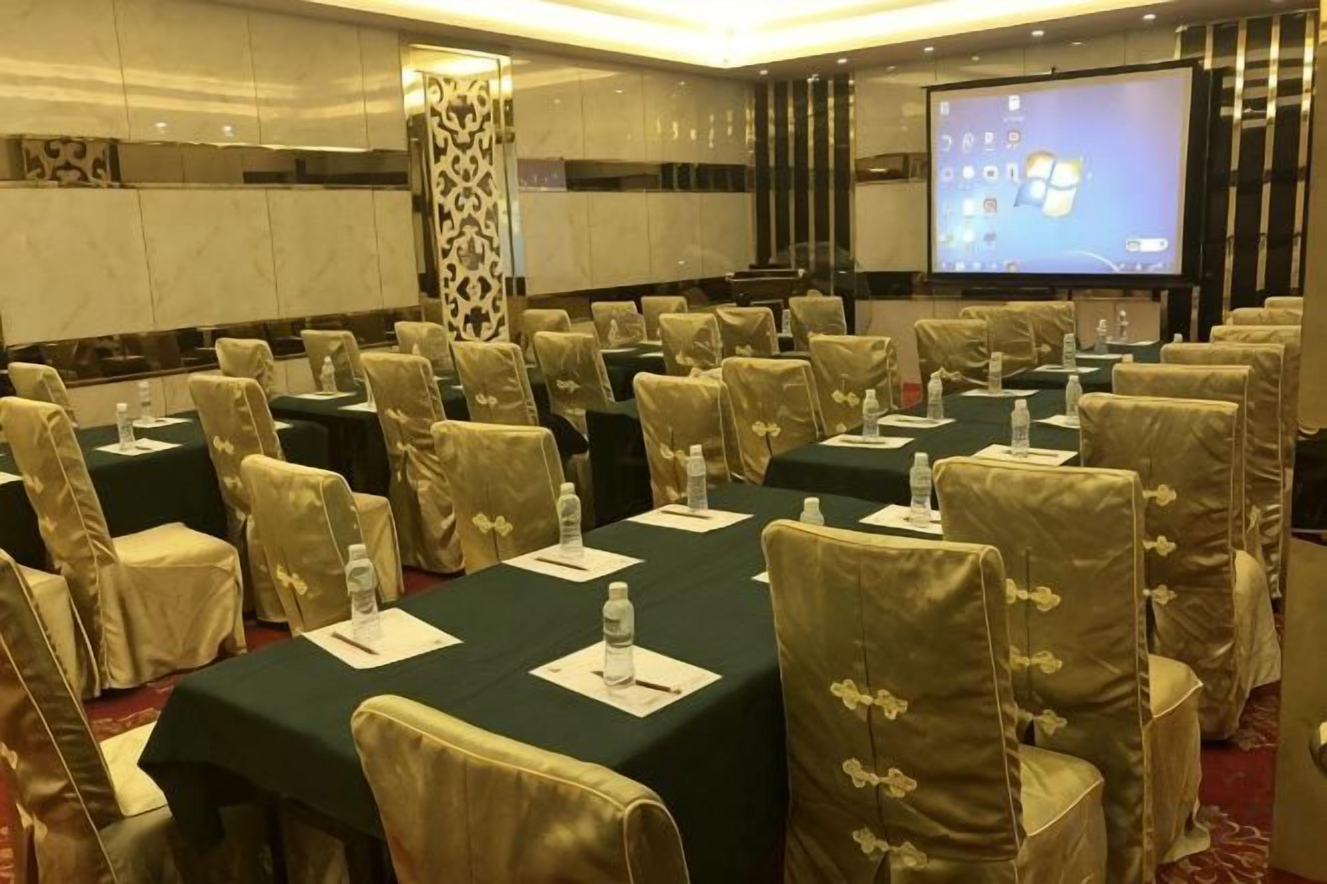 A conference room with rows of tables and chairs, a projector screen, and a large screen displaying a presentation.