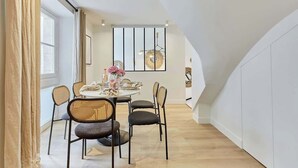 Un comedor con una mesa redonda dispuesta para cuatro personas, rodeada de sillas de mimbre. Se ve una ventana grande con cortinas a la izquierda, y una obra de arte colgada en la pared al fondo.