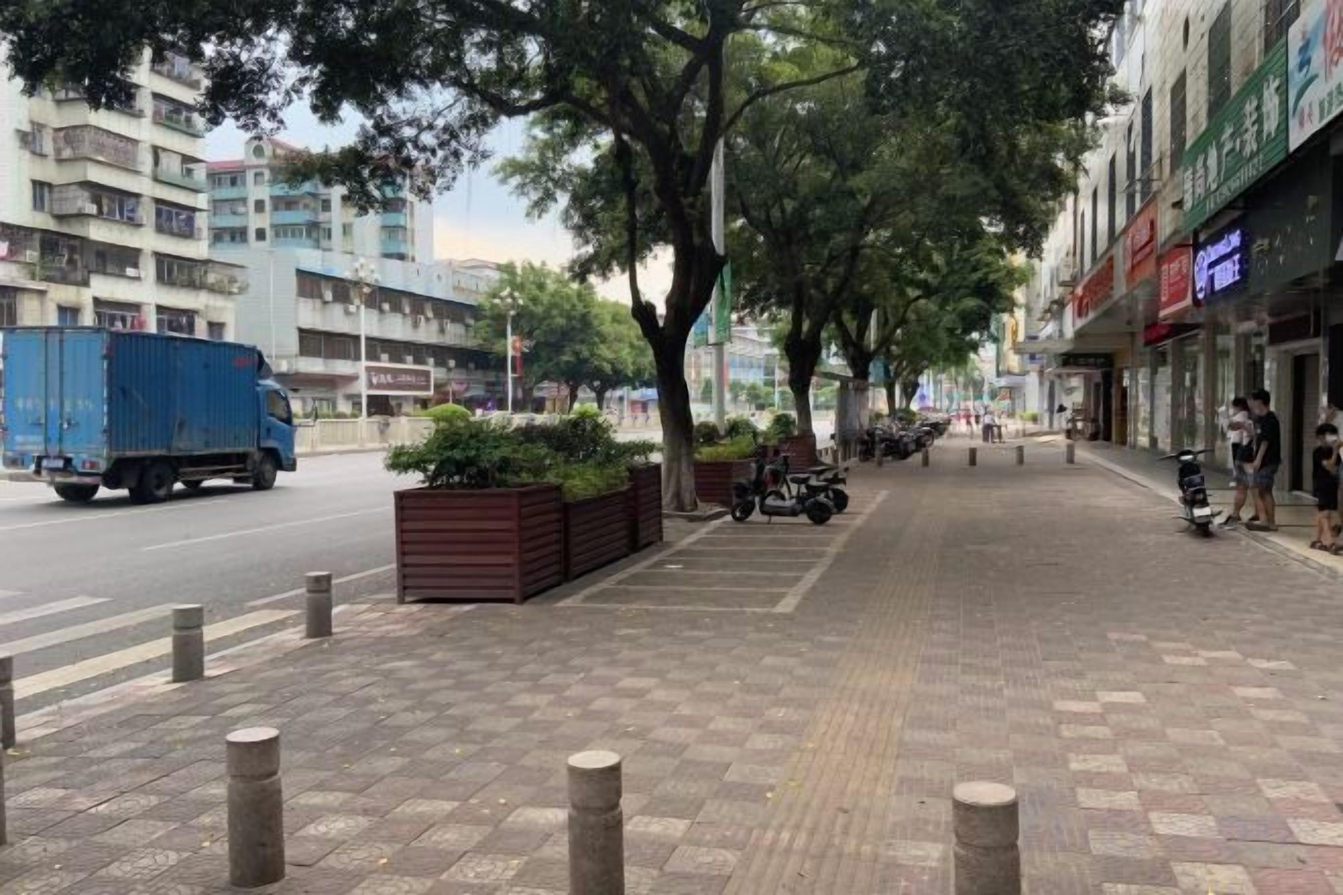Una acera con separadores, un camión azul en la calle y edificios con letreros.