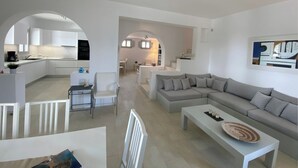 Ein modernes Wohnzimmer mit einem grauen Ecksofa, einem Couchtisch und einem Essbereich mit weißem Tisch und Stühlen. Im Hintergrund ist eine Küche mit weißen Schränken und einem schwarzen Backofen erkennbar.