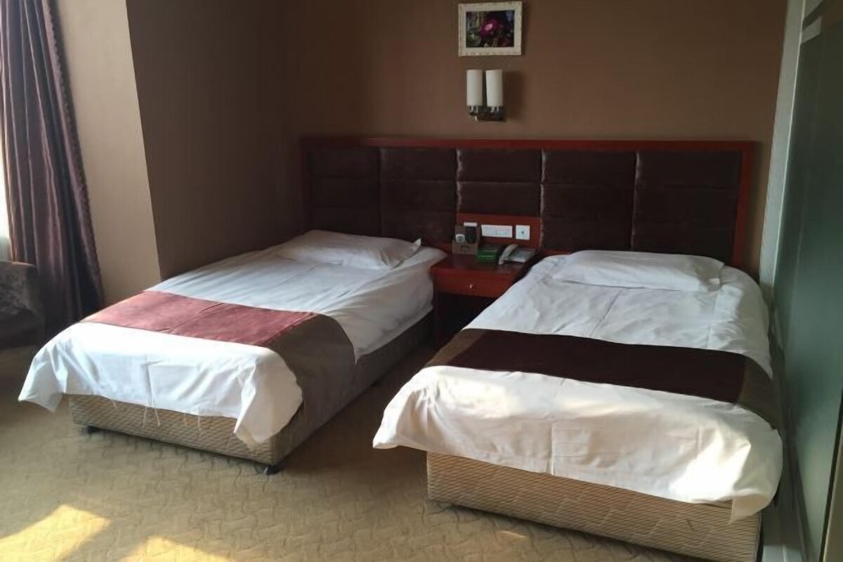 Een hotelkamer met twee eenpersoonsbedden, een houten hoofdeinde, een klein nachtkastje met een telefoon en een fles, en een wandlamp.