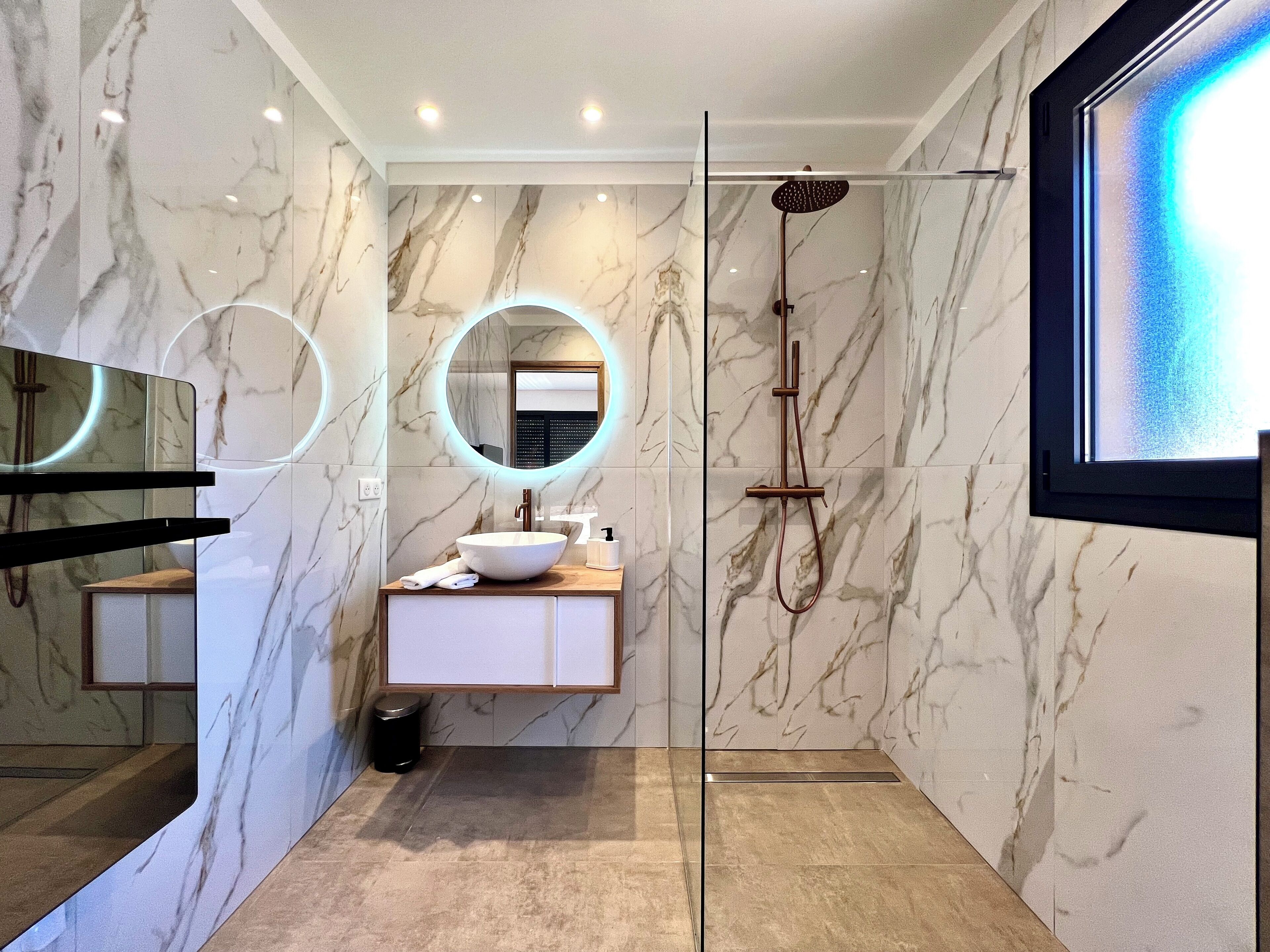 A modern bathroom with a marble wall, a round mirror, a wooden vanity, and a glass-enclosed shower.