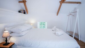 A bedroom with a wooden ceiling, a bed with white bedding, a bedside lamp, a small table, and a window with a view of greenery. - Briardene - 2 Bedroom Cottage - Oxwich (Swansea)