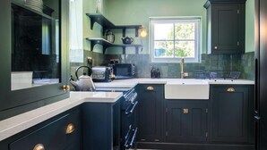 A kitchen with dark cabinetry, a white countertop, a window with a view of greenery, and a built-in oven. - Briardene - 2 Bedroom Cottage - Oxwich (Swansea)