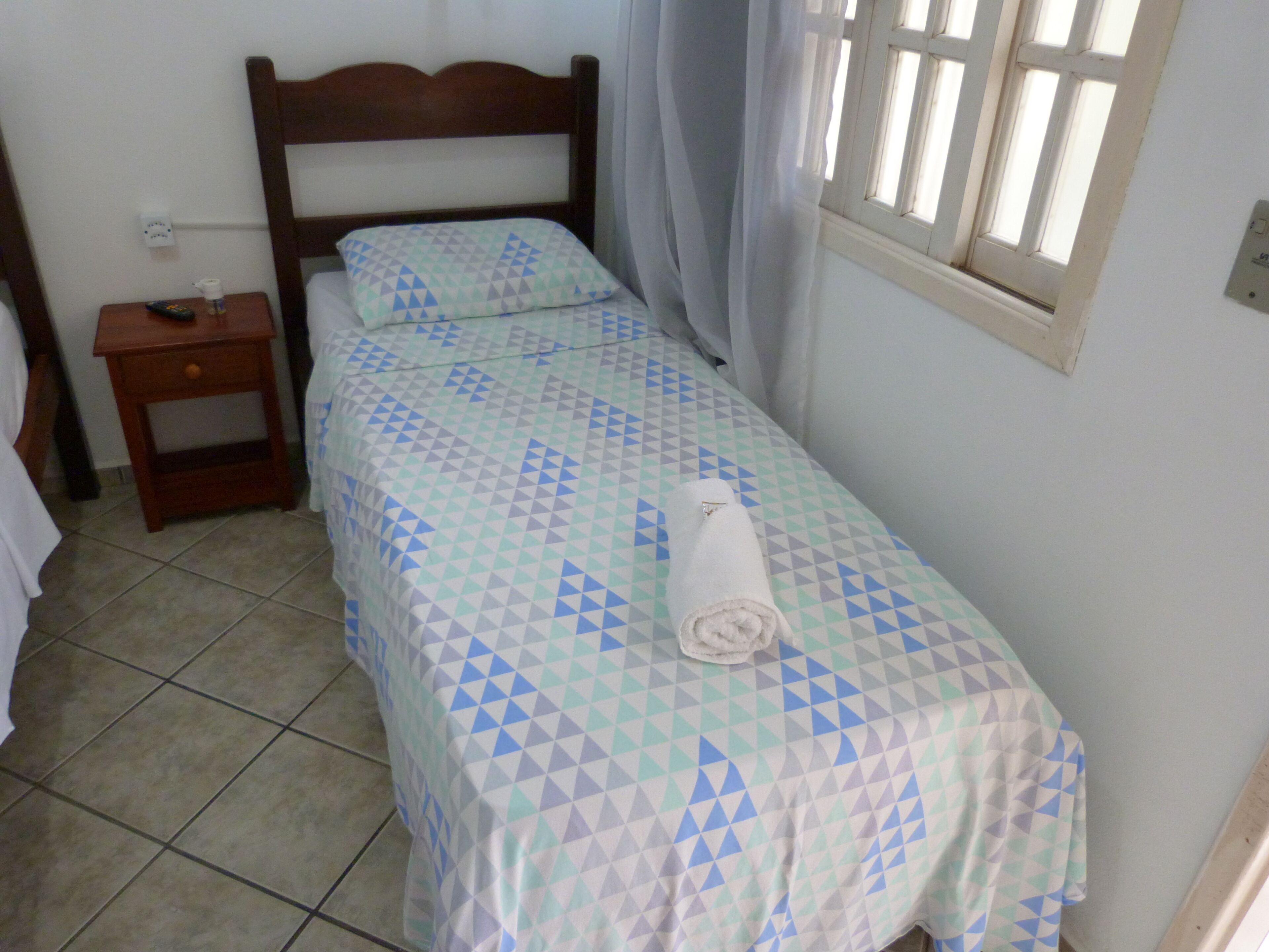 A single bed with a patterned bedspread, a wooden headboard, a nightstand with a remote control, and a window with curtains.