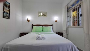 Un lit bien fait, avec du linge de lit blanc et des coussins verts, deux tables de chevet, un tableau encadré au mur et une fenêtre avec des rideaux.
