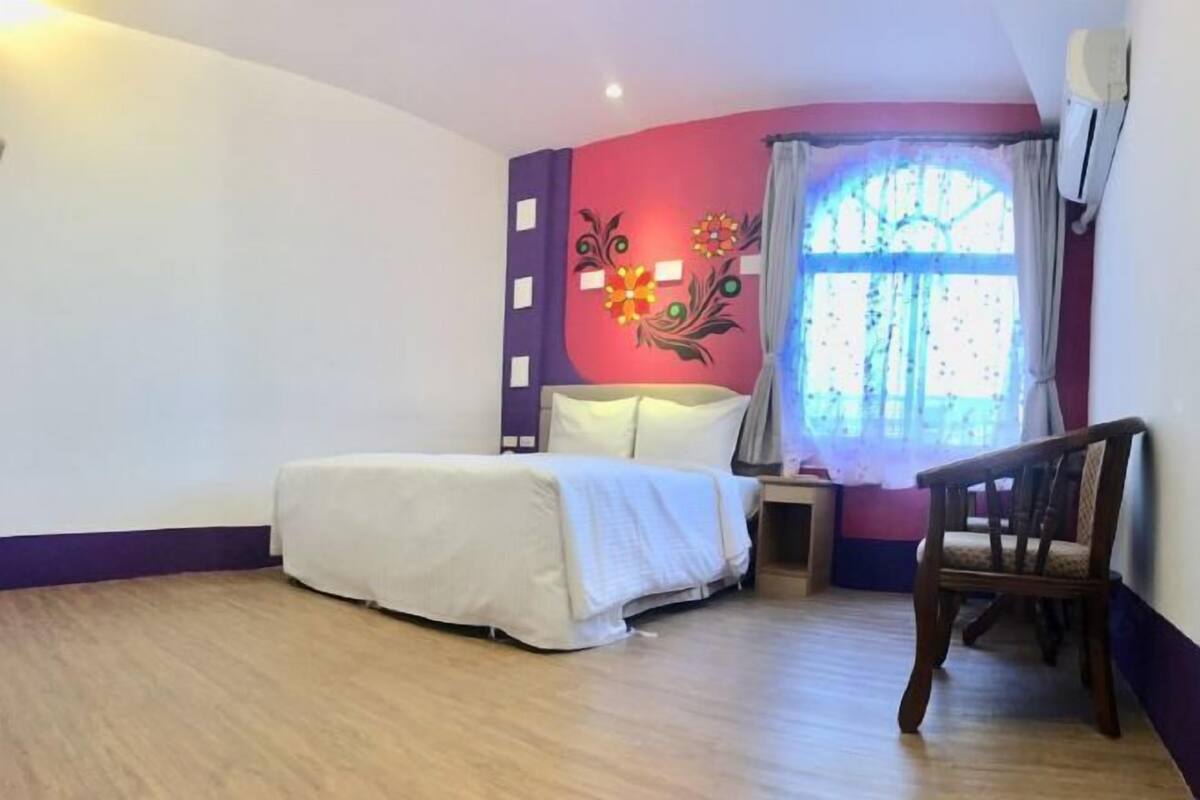 A bedroom with a bed, a chair, a nightstand, a wall with decorative elements, and a window with curtains.