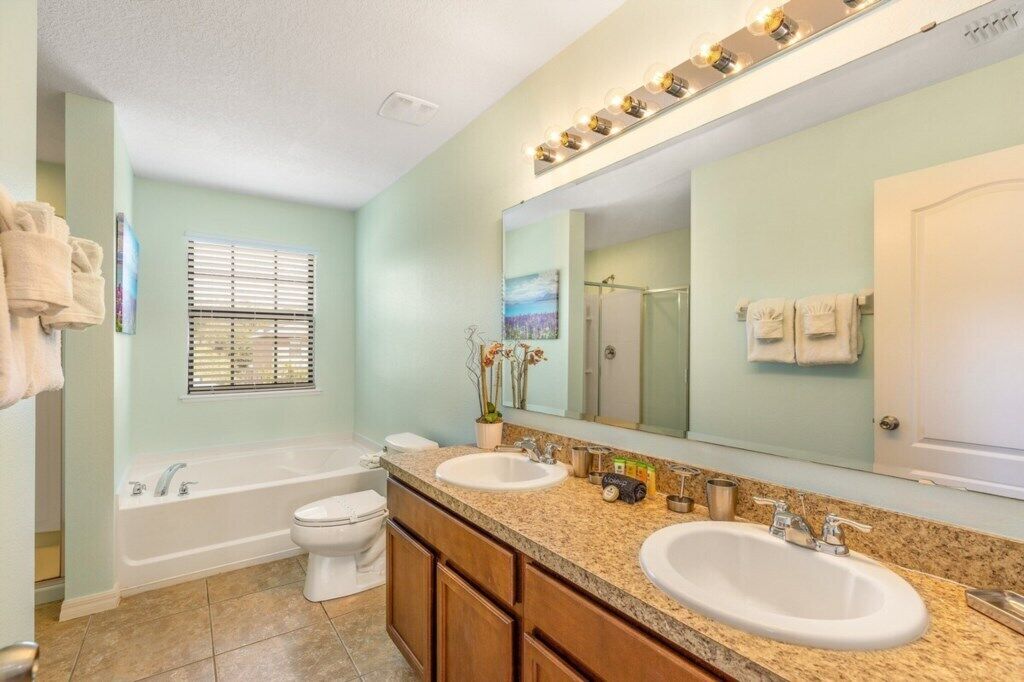 Une salle de bain avec deux lavabos, une baignoire et des toilettes. On y trouve une fenêtre avec des stores, un miroir avec éclairage intégré et un porte-serviettes.