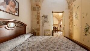 A bedroom with a wooden headboard, a floral bedspread, stone walls, and a view into another room with a dining table.