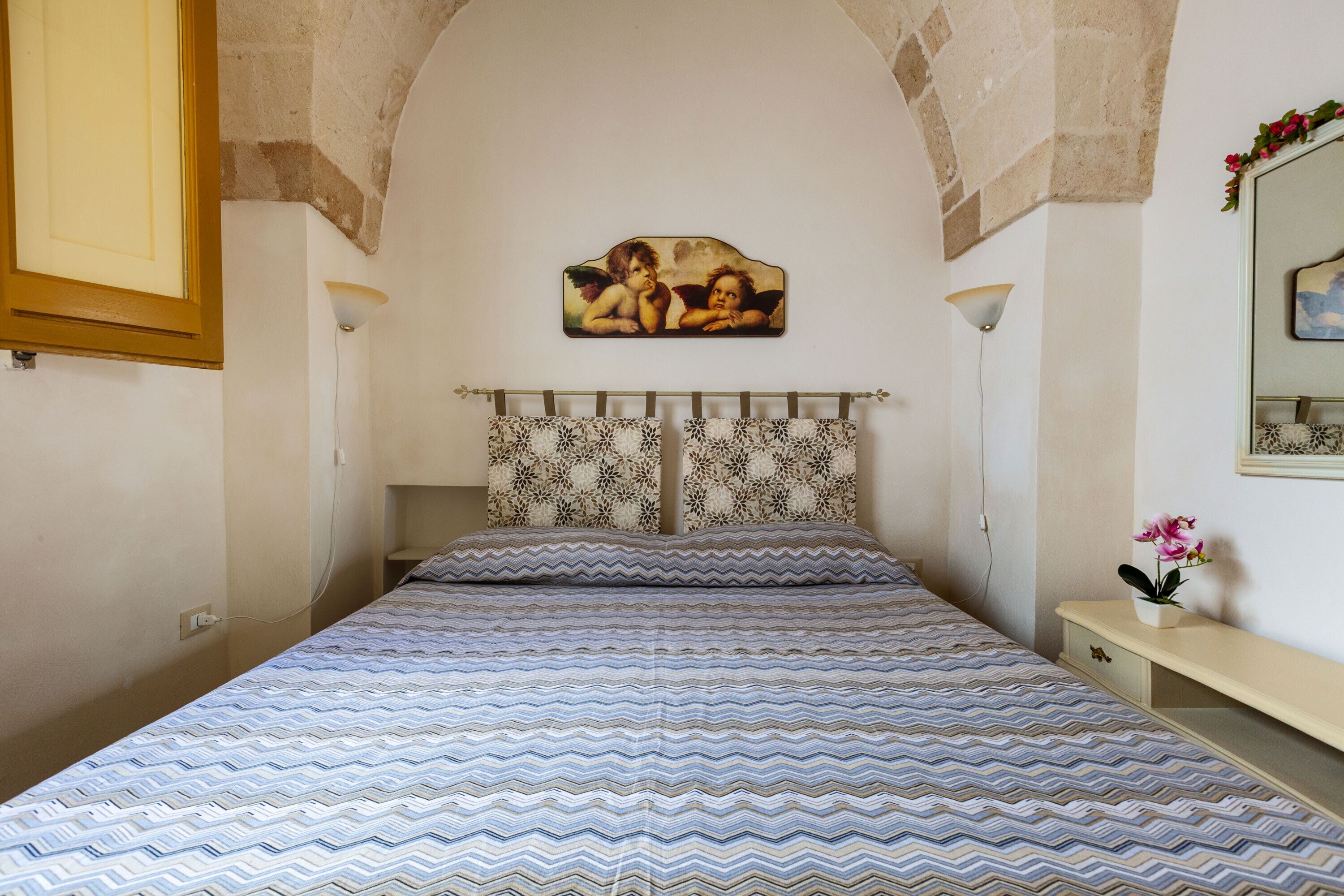 Une chambre à coucher avec une couvre-lit à motifs, un tableau représentant des anges sur le mur et une petite table de chevet agrémentée d’une plante.