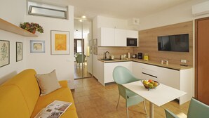 A compact living space with a kitchenette, a dining area with a table and chairs, a sofa, and a TV mounted on the wall. - I Girasoli D10 by Wonderful Italy (Toscolano Maderno)