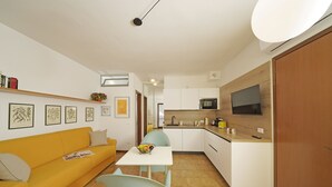 A compact living space with a yellow sofa, a dining table with chairs, a kitchen area with a microwave, and a TV mounted on the wall. - I Girasoli D10 by Wonderful Italy (Toscolano Maderno)