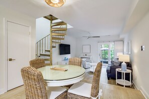 Un coin repas avec une table ronde en verre, des chaises en osier, un escalier en colimaçon et un salon comprenant un canapé et une lampe.