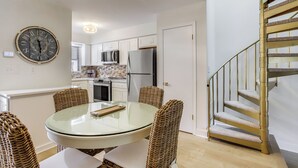 A modern kitchen with white cabinets, a stainless steel refrigerator, and a dining area with wicker chairs and a glass table. - Walk to Shore & Boardwalk: Bethany Beach Retreat (Bethany Beach)
