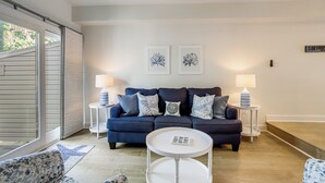 A modern living room with a dark blue sofa, two side tables with lamps, a white coffee table, and a large window with blinds. - Walk to Shore & Boardwalk: Bethany Beach Retreat (Bethany Beach)