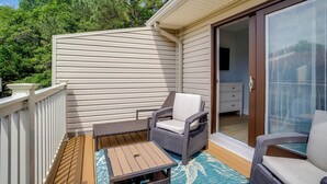 A covered patio with a wooden deck, two wicker chairs, a small table, and a sliding glass door leading to a bedroom. - Walk to Shore & Boardwalk: Bethany Beach Retreat (Bethany Beach)