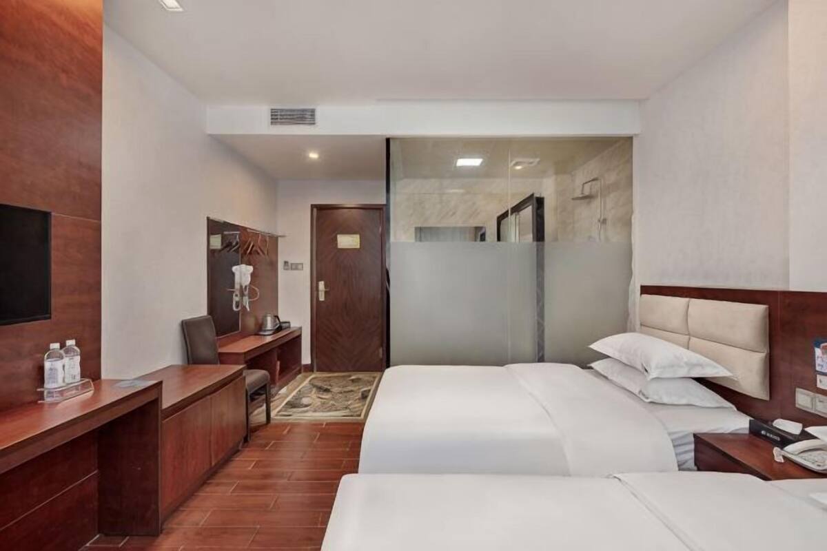 A hotel room with two beds, a desk, a chair, a TV, and a bathroom visible through a glass door.