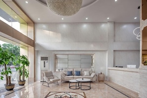 Un vestíbulo amplio con techo alto, un candelabro, una zona de estar con sofá y sillones, un mostrador de recepción y un ventanal grande.