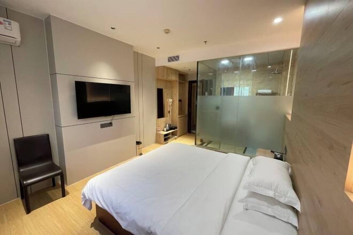 A modern hotel room with a large bed, a flat-screen TV, a chair, and a glass shower enclosure.