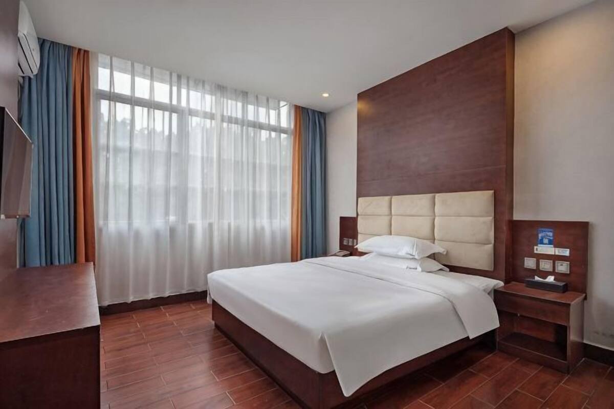 A hotel room with a large bed, a wooden headboard, a bedside table, a TV, and a window with curtains.