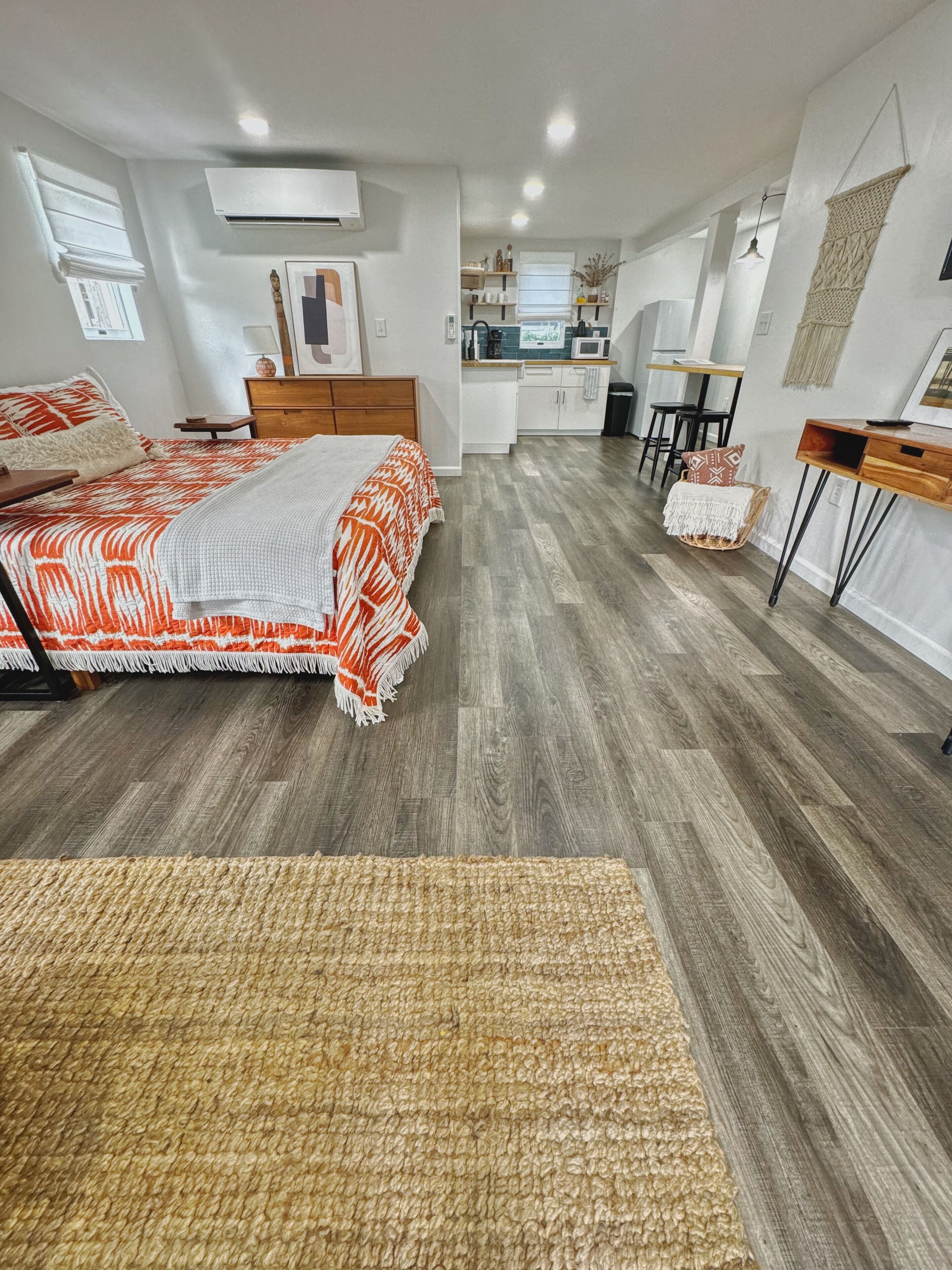 A modern bedroom with a large bed, a wooden floor, and a kitchenette in the background.