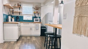 Une cuisine moderne avec des armoires blanches, un plan de travail en bois et une poubelle noire. On y trouve une chaise en osier, recouverte d’un plaid blanc et agrémentée d’un coussin à motifs.