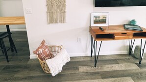 Un salon moderne avec un bureau en bois, une décoration murale en macramé tissé, un fauteuil en osier agrémenté d’un plaid blanc et un téléviseur à écran plat fixé au mur.