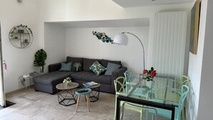 A modern living room with a sofa, two side tables, a glass dining table with chairs, a wall-mounted lamp, and a decorative wall piece. - Pretty harbour house with garden (Mandelieu-la-Napoule)