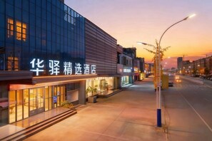 A hotel with a modern facade, illuminated signage, and a wide street in the background. - Home Inn Alliance Huayi Hotel (Tancheng Railway Station Tanguo Ancient City) (Tancheng)