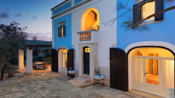 A courtyard with a stone floor, a dining area with a table and chairs, and a building with a blue facade and arched windows.