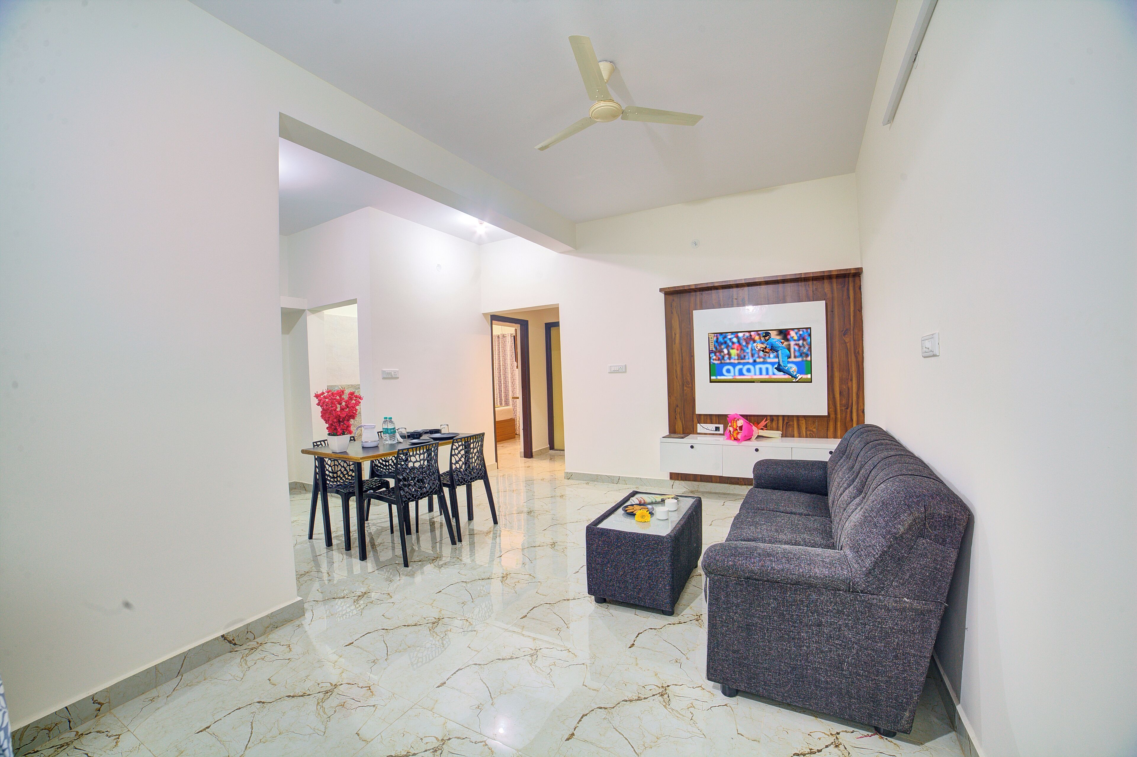 A living room with a sofa, a dining table with chairs, a TV mounted on the wall, and a ceiling fan.