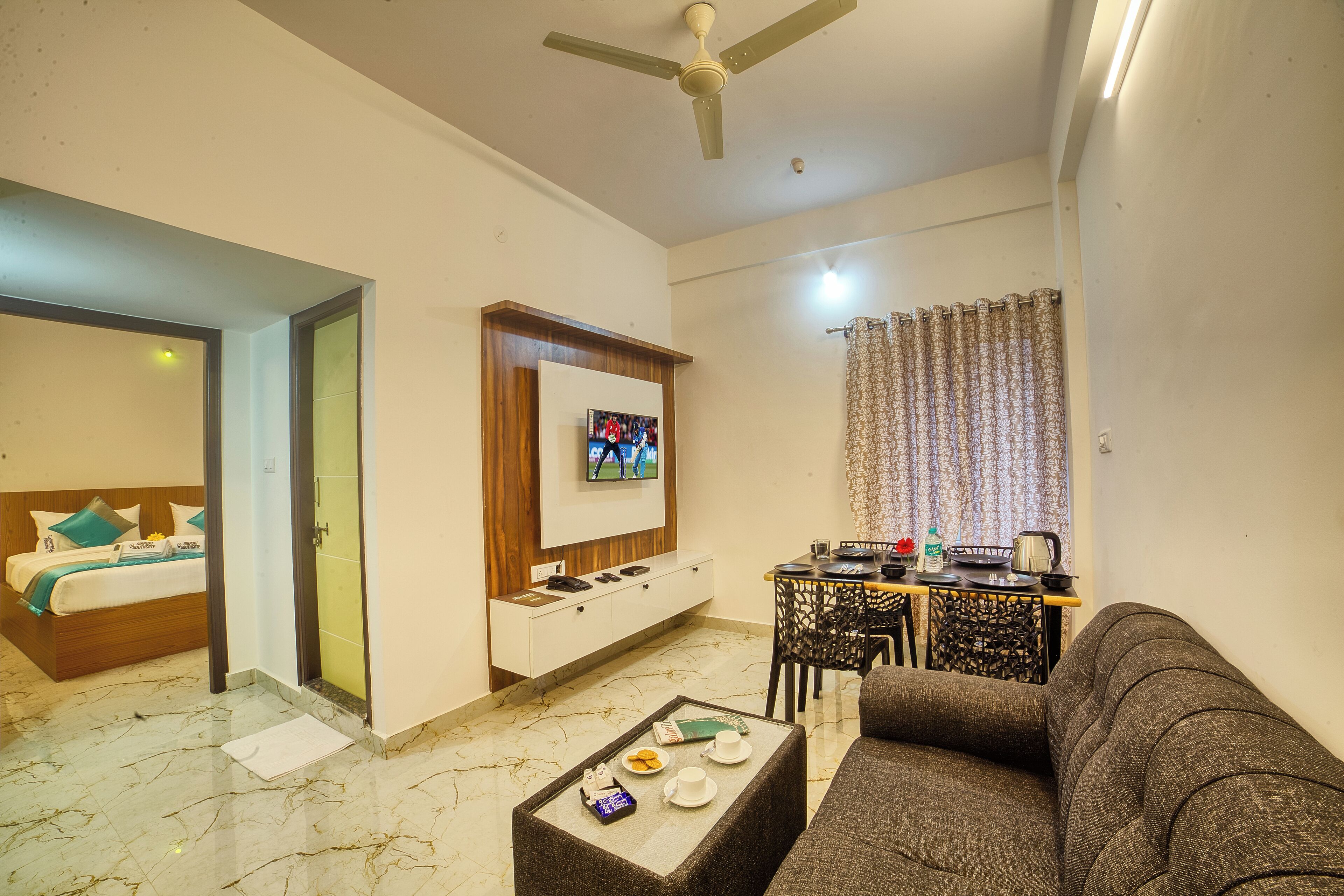 A modern living room with a sofa, a coffee table, a TV mounted on the wall, a dining area with a table and chairs, and a bed with a wooden headboard.