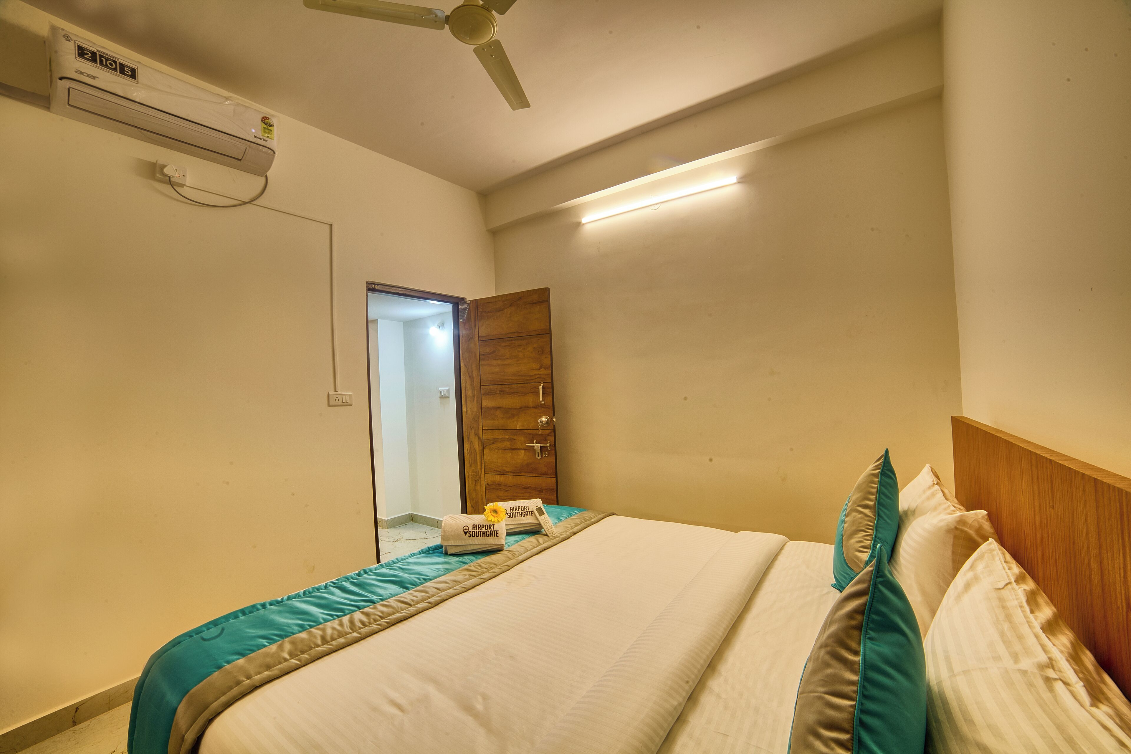A hotel room with a bed, a ceiling fan, an air conditioning unit, and a wooden door.