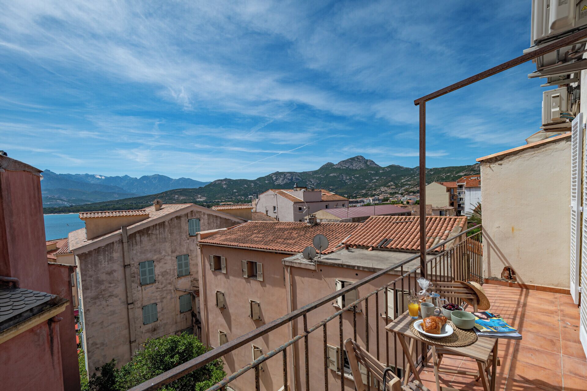 Una terraza con una mesa preparada para el desayuno, con vistas a un pueblo costero de edificios tradicionales y una montaña al fondo.