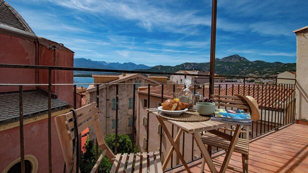 Un balcon avec une table préparée pour le petit-déjeuner, offrant une vue sur un paysage montagneux et une étendue d’eau.