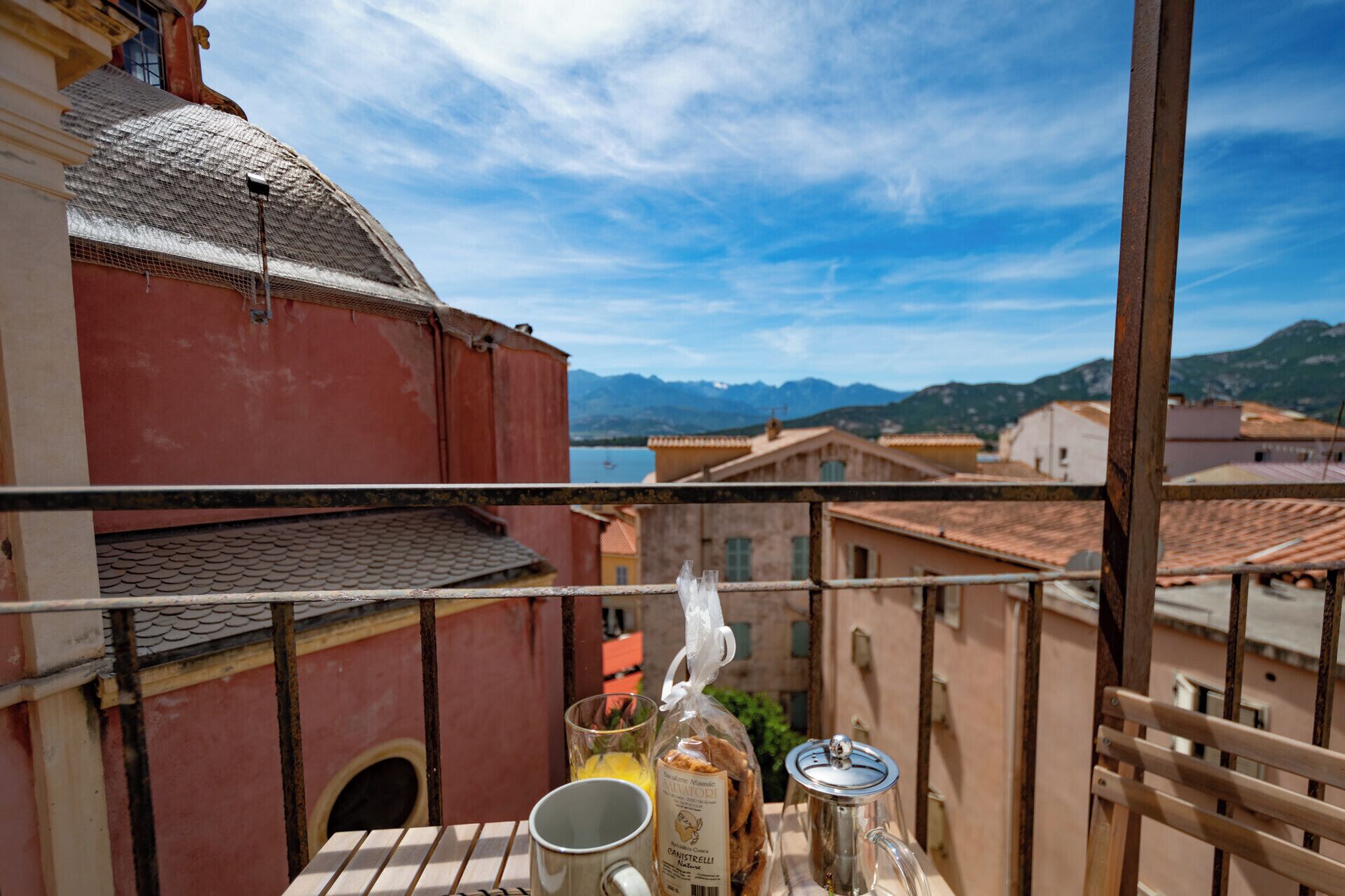 A balcony with a view of a red building, a mountain range, and a clear sky.