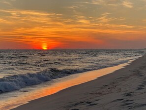 A beach with a sunset over the ocean, waves crashing onto the shore, and a clear sky.