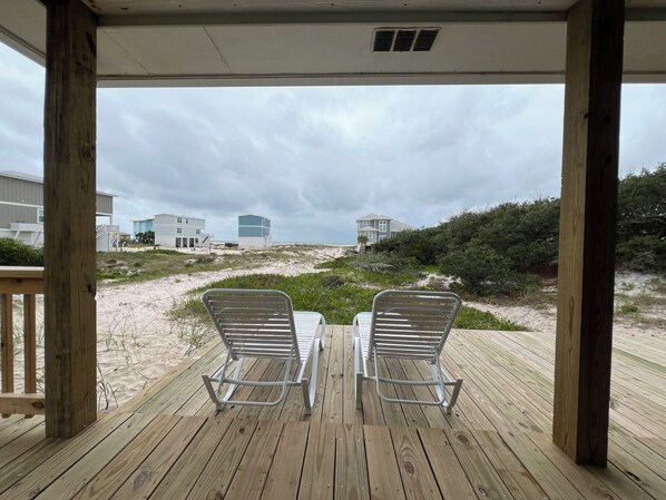 A wooden deck with two metal chairs facing a beach with buildings in the background.