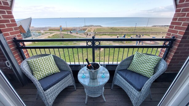 A balcony with wicker chairs, a small table with a bottle and glasses, and a view of the beach and ocean.