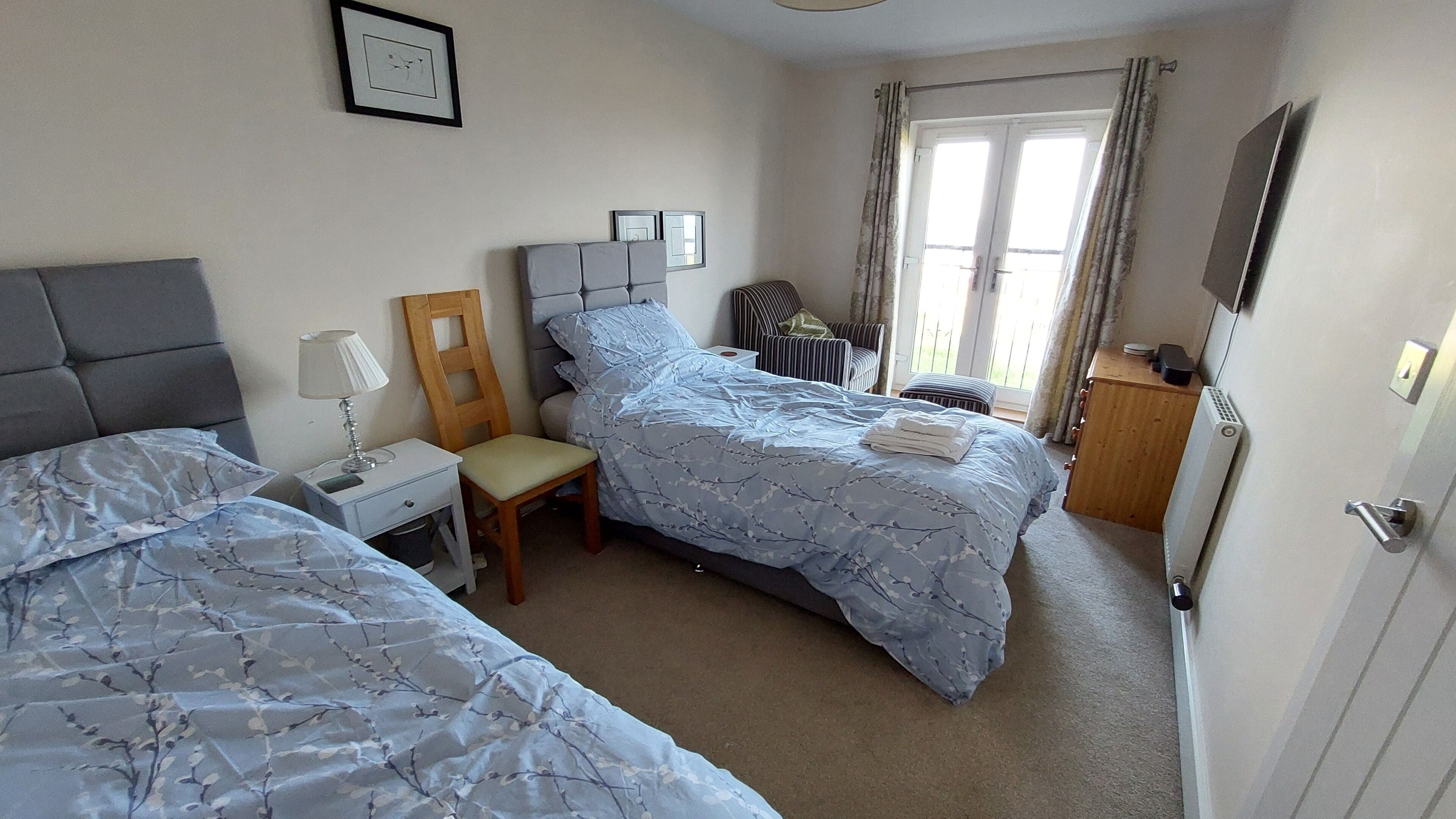 Quarto com duas camas, uma cadeira, mesa de cabeceira com abajur, radiador e janela com cortinas.