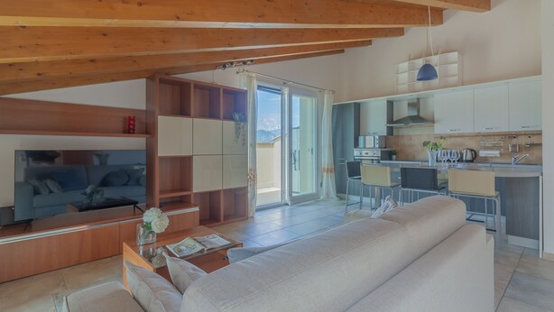 A modern living room with a large flat-screen TV, a sofa, and a coffee table. There is a sliding glass door leading to a balcony with a view of the mountains. The kitchen is visible in the background with white cabinets and a blue pendant light. - Magic Lake View in Lierna (Lierna)